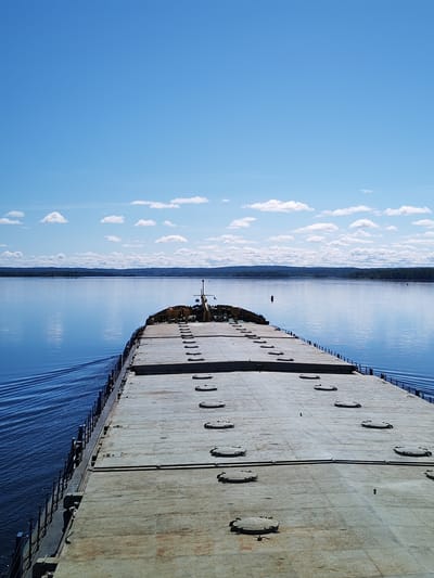 This image presents a serene daytime view from the deck of a large vessel, likely a barge or cargo ship, navigating a wide body of water near Shestovo, Russia. The perspective is looking forward over the vessel's long, flat deck towards the horizon.
The foreground is dominated by the ship's deck, a broad, weathered expanse of gray or off-white metal. Numerous circular hatches, possibly covers for cargo holds, are evenly spaced across the deck. Towards the bow, some industrial equipment or a small superstructure is partially visible, along with railings lining the sides of the ship.
To the left of the vessel, a distinct wake trails across the calm, deep blue water, indicating the ship is moving forward. The water beautifully reflects the bright blue sky above, creating a seamless blend of blues. A single red buoy can be seen floating in the water in the middle distance, to the right.
In the far distance, a low, dark green tree-lined horizon stretches across the frame, marking a distant shoreline or landmass. The sky above is a clear, vibrant blue, adorned with scattered, fluffy white cumulus clouds.
The scene conveys a sense of peaceful travel under clear, sunny weather conditions, likely mid-morning or afternoon given the bright illumination. No people or visible text are present in the image.