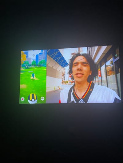 A person wearing an Adidas shirt with a black, red, and gold collar is holding a phone. The phone screen displays a selfie video with a Pokemon Go screen at the bottom. The Pokemon Go screen shows a Cyndaquil. The background of the selfie appears to be an urban setting with buildings. The image was likely taken in the city of Saedinenie, Bulgaria.