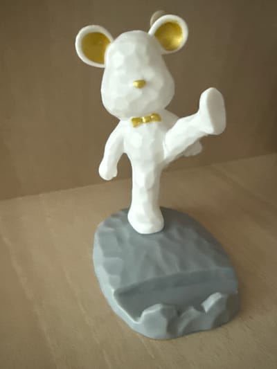 A white, faceted figurine of a mouse stands on a gray, rocky base. The mouse has large, circular ears with gold interiors and a small gold bow tie. It is posed with its right leg extended backward and its left leg bent. The background is a plain, light brown surface, likely a wall or shelf. The base of the figurine is on a light brown, wooden surface, possibly a table or shelf. The overall scene is well-lit, and the focus is on the figurine. There is no information available about the location being in Hraniv, Ukraine.