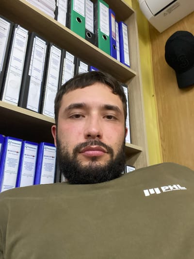 The image shows a medium close-up selfie of a young man with a beard and short dark hair, taken indoors in what appears to be an office. He is wearing a dark olive green crewneck sweatshirt with a white logo on the left chest that reads "PHL" with smaller text below that is unreadable. The background consists of several shelves filled with blue and white binders. To the right of the shelves, a light yellow wall is visible, and on the upper right, part of a white air conditioner and a black baseball cap hanging on the wall can be seen. The scene is consistent with a typical office environment in Luzhitsy, Russia.