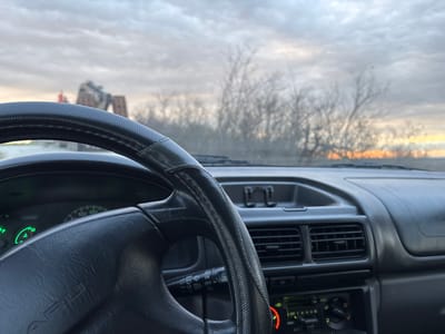 A view from the interior of a Subaru car, focusing on the steering wheel and dashboard, with an external view visible through the windshield. Outside, there are bare trees in the background and construction equipment is present. The sky is overcast. The location is speculated to be in Gornik, Bulgaria.