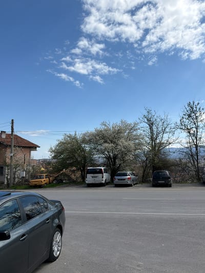 The image shows a street scene in Sveti Konstantin, Bulgaria. The view includes a portion of a building and utility pole at the bottom. Parked parallel along the edge of the road are five cars, with a grey sedan in the immediate foreground. Behind the grey car are two white vans, a silver car, and a black van. Trees are situated between the parked cars and a glimpse of buildings and scenery in the distance. The sky, which occupies a significant portion of the image, is blue with scattered white clouds. The road is asphalt with painted white lines. The photograph appears to have been taken during the day, under natural lighting conditions.