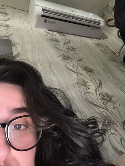 The image shows a partial view of a person with dark hair and round glasses. Only one eye is visible. The person appears to be lying down on a surface covered with a patterned fabric. An air conditioning unit is mounted on the wall next to the person. The location is not identifiable as Dobrovnitsa, Bulgaria from this image.