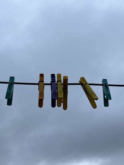 The image shows a close-up, low-angle view of six colorful plastic clothespins attached to a thin, black clothesline. The clothespins are arranged from left to right: one green, two yellow, one purple, one yellow, and one green. The background is a cloudy, overcast sky. The scene is set in Gonja, Russia. There are no people or actions depicted.