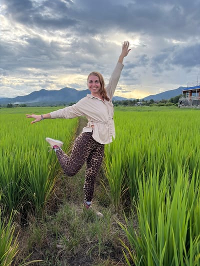 In the city of Ban Pong, Thailand, a woman with blonde hair is captured in mid-air, posing in a vibrant green rice paddy. She is wearing a light-colored, loose-fitting top and leopard-print leggings, with her right leg bent at the knee and her left leg extended behind her. Her right arm is raised, and her left arm is outstretched to her side. The sun is setting, casting a warm glow over the scene, with mountains visible in the background and a building with a blue roof to the right. The sky is filled with dramatic clouds.