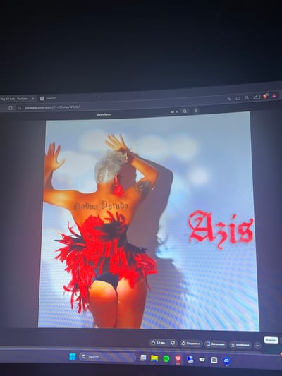 The image shows a person with short, white hair, facing away from the camera. They are wearing a black bikini bottom and a red feathered headdress or garment around their waist and chest. The person has tattoos on their back and arms. There is text on their back that reads "Sadna Poroda" in gothic lettering, and red text on the right side of the image that reads "Azis" in a stylized font. The background is a light blue, possibly a wall or a sky. The image appears to be a screenshot from a YouTube video, as indicated by the browser window elements at the top and bottom of the image. The location is not specified in the image.