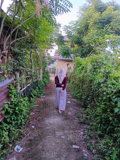 A person wearing a light brown hijab and a maroon long-sleeved shirt stands with their back to the camera, facing away down a narrow, unpaved alleyway. They are wearing wide-striped, light-colored pants and are holding their hands clasped behind their back. The alley is flanked by lush green foliage on both sides, with some trees whose branches extend over the path. On the left side of the alley, a corrugated metal fence can be seen partially obscured by vegetation. In the background, buildings with red-tiled roofs are visible through the trees. The ground of the alley is a mixture of dirt, gravel, and fallen leaves, with some scattered debris. The lighting suggests it is daytime, possibly late afternoon or early morning, with soft light illuminating the scene. The overall environment appears to be a semi-rural or residential area within the city of Meuliek, Indonesia, characterized by dense vegetation and simple structures.