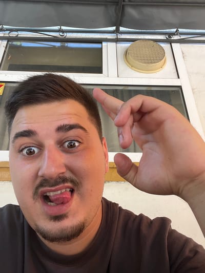 A close-up, waist-up, selfie-style photograph features a young man with dark hair, a mustache, and a goatee. He is wearing a dark brown t-shirt and is positioned in the foreground. His facial expression is animated, with wide-open eyes and his tongue playfully extended. His right hand is raised towards his head, with his index finger extended and bent as if making a gesture.
Behind him, a window frame is visible, with panes that reflect the sky and surrounding environment. Above the window, a grey awning or overhang is present. To the right of the window, a round, textured, light-colored object, possibly a vent cover, is mounted on the wall. The wall itself appears to be painted white and has a yellowish trim along the bottom.
The overall setting suggests an outdoor or semi-outdoor location, likely a public space given the presence of the window and exterior elements. The man's playful demeanor and the selfie format indicate a casual and personal moment. The image does not contain any specific landmarks or elements that would definitively place it in the city of Akandzhievo, Bulgaria.
