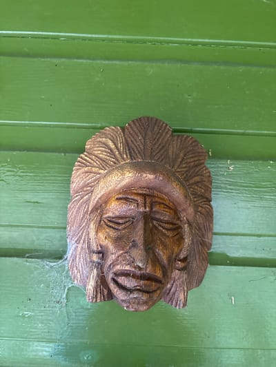A carved wooden mask, depicting a stylized human face with closed eyes, a prominent nose, and an open, grinning mouth, is affixed to a vibrant green, horizontally-paneled wooden surface. The mask is dark brown, exhibiting visible carving textures, and features a headdress resembling feathers. A white cobweb is present on the lower left side of the mask, extending onto the green background. The scene is located in the city of Stiperstones, United Kingdom.