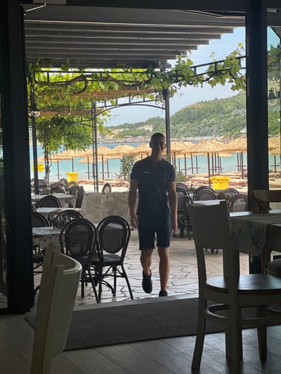 A young man wearing a dark polo shirt and shorts stands in the doorway of a restaurant, facing the camera with a smile. He is positioned in front of a row of tables and chairs that are set up for dining. Beyond the restaurant's open doors, a beach scene is visible with thatched umbrellas, lounge chairs, and the sea. In the background, a lush, green hillside overlooks the water. The restaurant's outdoor seating area is covered by a canopy of grapevines, which adds a rustic charm to the setting. The man appears to be a server or employee, given his attire and the location. The overall atmosphere suggests a relaxed, seaside dining experience in Topola, Bulgaria.