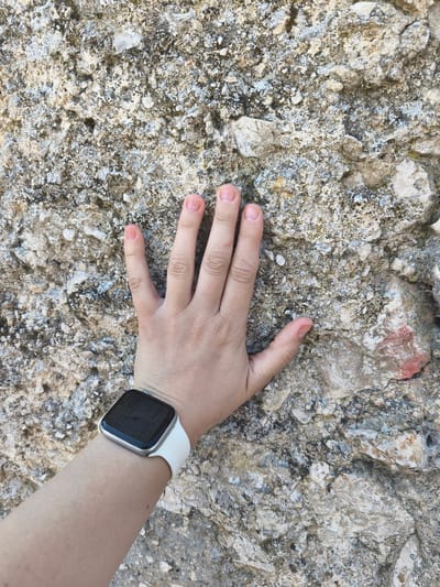 A close-up, high-angle shot shows a person's left hand with fingers splayed against a rough, textured rock surface. The hand is fair-skinned with short, light-colored fingernails. A white-strapped smartwatch is visible on the person's wrist, displaying a digital time of 10:09. The rock appears to be a light beige and gray, with some darker flecks and lighter white patches, possibly indicating mineral deposits or lichen. The texture of the rock is uneven and porous, with small pebbles and rough protrusions visible. The scene is outdoors, with natural lighting. The context of Campochiaro, Italy, is not visually discernible from the image alone.
