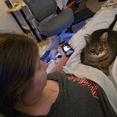 The image depicts an indoor scene, likely a bedroom. A person, seen from behind and to the side, is lying on a bed and looking at a smartphone. The person has brown hair and is wearing a grey t-shirt with a necklace. Next to the person on the bed is a tabby cat, lying down and looking towards the camera. The bed is covered with a duvet featuring a white polka dot pattern. On the left side of the image, a blue object with a picture on it is visible on the bed. In the background, there is a desk with items on it, including what appears to be a computer monitor and an office chair. The overall lighting suggests an evening or dimly lit environment. The image does not provide any specific visual cues to determine its location within Portland, United States of America.