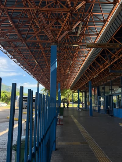 The image captures an expansive, outdoor public transport station or sheltered area, likely located in Bjeliši, Montenegro, under bright daylight. A dominant feature is a massive, rust-colored or dark orange metal truss structure forming an extensive canopy or roof overhead. This intricate framework of interlocking triangular beams supports a roof made of dark grey corrugated panels. Several prominent blue columns provide additional support for the structure.
Parallel to the roof, on the right side of the frame, runs a long, blue metal fence or railing, designed to guide pedestrians or separate areas within the station. The ground beneath is paved with dark grey interlocking bricks, featuring a distinctive yellow tactile paving strip that stretches into the background, intended to assist the visually impaired.
In the mid-ground and background, beyond the blue fence and some smaller blue structures (possibly kiosks or bus shelters), a few blurry human figures are visible. One figure appears to be pushing a cart. The faint outlines of buses or other large vehicles can be seen further back, affirming the setting as a transportation hub. A small white car is also partially visible in the far distance.
The sky is clear and blue with scattered white clouds, indicating sunny weather. Lush green foliage from trees and bushes frames the background, and the gentle contours of hills or mountains are discernible on the horizon. A white sign with the black number "5" is visible in the distance, likely indicating a bus bay or platform. The scene depicts a functional and well-structured public transport facility.