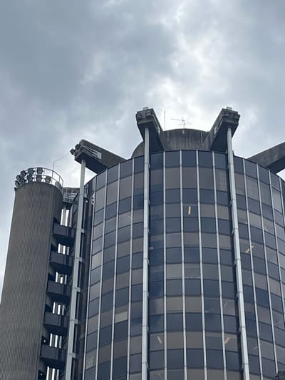 The image is a low-angle, vertically oriented shot of a modern building with a glass facade, set against an overcast sky. The building's architecture is characterized by sharp, angular protrusions and horizontal bands of windows. Some windows appear to be lit from within, suggesting it's either daytime with interior lighting or dusk/night. The building has a ribbed cylindrical section at the bottom, topped with a ring of spotlights. The sky is filled with dense, grey clouds, indicating a cloudy or possibly rainy day. There is no visible text in the image. The scene appears to be an urban environment, likely in Créteil, France, given the location context. No people are visible in the frame.