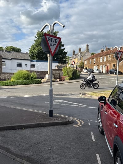 This image captures a detailed street scene at an intersection, characteristic of a town or village in the United Kingdom, specifically identified as King's Lynn and West Norfolk.
In the immediate foreground on the right, the front-right quarter of a red Mini Cooper Countryman is visible, positioned alongside the road with its alloy wheel and black fender trim prominent. The asphalt road surface shows dashed white lines and double yellow lines near the kerb.
A tall, grey metal street pole stands centrally in the foreground, topped with two curved silver arms. Attached to this pole is a prominent triangular "GIVE WAY" sign, featuring a red border, white background, and black capitalized text.
In the midground, a motorcyclist is actively navigating the intersection on a dark-colored street motorcycle. The rider is wearing a grey full-face helmet, a light-colored long-sleeved top, dark trousers, and a black backpack. The motorcycle is equipped with disc brakes and a rear mudguard. The road surface at the intersection is marked with dashed white lines. Further down the street to the left, a second, smaller "GIVE WAY" sign is visible.
The background reveals a mix of architecture and public amenities. On the left, a white single-story building with multiple dark-framed windows and a dark roof is partially obscured by trees. In front of this, a well-maintained public area features a low stone wall, manicured green hedges, bushes, and a distinct stone monument, possibly a war memorial, with stone steps and two wooden benches positioned on either side. Beyond this, a row of traditional British terraced houses line the street. These buildings display varying shades of brick and stone, multiple windows, pitched tiled roofs, and prominent chimneys. Several parked cars, including light-colored hatchbacks and a dark sedan, are visible along this street.
The sky above is overcast with a mix of grey and brighter cloud formations, indicating a partly cloudy day.