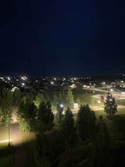 The image displays an aerial view of a small town or village at night. The scene is dominated by the dark night sky on the left, with a few distant, faint lights visible. On the right, a suburban area is illuminated by numerous streetlights, casting a yellow glow on the paved roads, sidewalks, and grassy areas. Several houses with lit windows are scattered throughout the illuminated section, suggesting a residential neighborhood. Tall trees with dense foliage are interspersed with the buildings, their leaves catching the light and creating shadows.
The time of day is clearly night, as evidenced by the darkness of the sky and the artificial lighting. There are no visible weather conditions, but the air appears still. The overall atmosphere is peaceful and quiet, typical of a residential area after dark. The location cues suggest a somewhat rural or suburban setting, with a clear distinction between the undeveloped, dark expanse and the developed, lit up area. No people are visible in the frame, and the objects are primarily houses, streetlights, trees, and paved pathways. There is no visible text in the image.