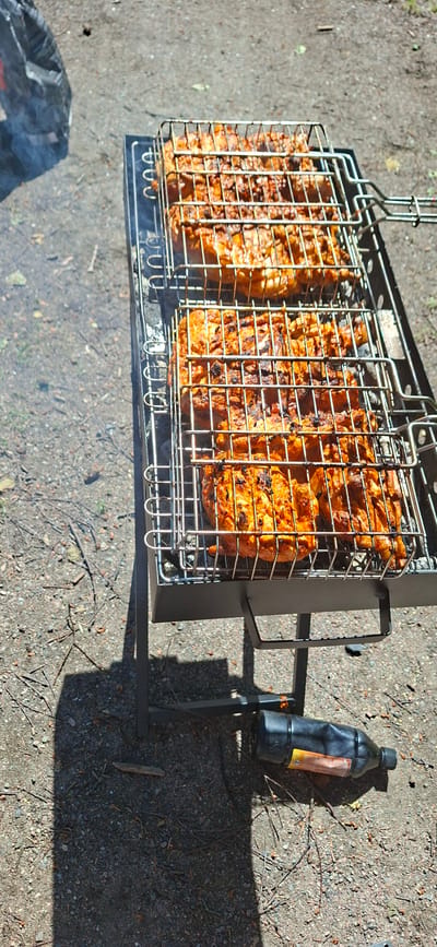 In the city of Lahti, Finland, outdoors on a gravelly surface, a portable charcoal grill is set up. The grill holds two metal baskets filled with seasoned meat, likely chicken, which is actively cooking over charcoal. Smoke rises from the grill, indicating it is in use. To the left of the grill, a black plastic bag, possibly containing charcoal, is visible. On the ground beneath the grill, a black plastic bottle with an orange and yellow label lies on its side. The scene is bathed in sunlight, casting shadows on the ground.