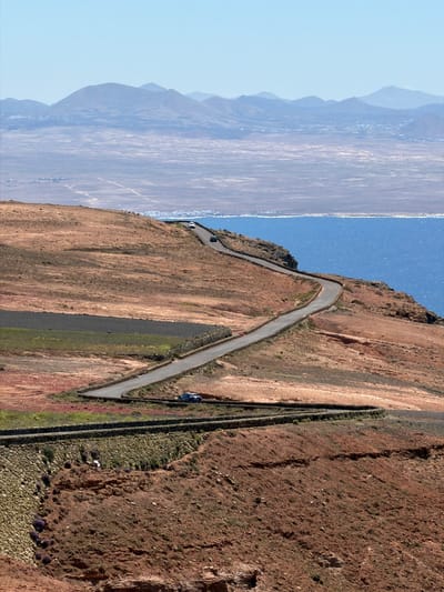 The image presents an aerial or high-vantage view of a winding road traversing a steep, arid hillside, overlooking a vast expanse of ocean and distant land.

**Subjects:**
The primary subject is a narrow, asphalt road, characterized by several sharp turns, snaking through the rugged, reddish-brown terrain. Two cars are visible: a white car is positioned higher up the road, and a darker vehicle (possibly blue) is on a lower section towards the right. The landscape is dominated by rocky, barren mountain slopes, interspersed with sparse green vegetation and patches of smaller loose rocks. A long, dark, linear feature, resembling a stone wall or track, runs parallel to the road on the right. In the foreground, clusters of purple flowering plants add a touch of color. A wide body of sparkling blue water, likely the ocean, occupies the mid-ground, beyond which faint, hazy silhouettes of distant mountains or landmasses are visible.

**Activity or Event:**
The scene captures a moment of travel or exploration along a scenic mountain road. The cars indicate ongoing vehicular movement or a stop along this route. No explicit human activity, such as hiking or direct interaction, is clearly depicted.

**Notable Details:**
The image is set during bright, clear daylight, likely late morning or early afternoon, with strong illumination and a cloudless, light blue sky. The weather is excellent. The rugged, volcanic-like landscape, winding coastal roads, and the presence of a vast ocean are strong location cues consistent with the provided context of Haría, Spain, characteristic of the Canary Islands. The perspective offers a panoramic view, emphasizing the dramatic scale of the natural environment. The color palette features earthy reds and browns, contrasting with the deep blues of the water and sky, and accents of green and purple vegetation.

**Visible Text:**
There is no visible text in the media file.