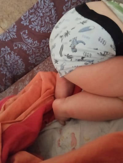 An overhead close-up shot captures the lower torso and bent legs of a baby or young child, likely resting on soft furnishings in Teykovo, Russia. The child is wearing a light-colored diaper or pull-up, which features a repeating pattern of surfboards, palm trees, and the text "SURF", "COOL DUDE", and "1983". A black elastic waistband is visible at the top of the diaper. The child's legs are bent, with their knees drawn upwards, indicating a relaxed posture, possibly sleeping or resting.

To the left of the child, a dark reddish-brown fabric with an intricate light blue or grey damask-like pattern is visible, likely a pillow or blanket. Below the child's waist, an orange and deep red layered fabric, possibly a blanket or towel, adds a warm color to the scene. The child's legs rest on a white sheet or blanket, which has some faint, irregular pink and yellow stains or patterns. The overall setting appears to be indoors, possibly on a bed or sofa, with no specific time of day or weather cues discernible from the lighting.