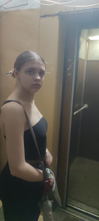 A young woman with light brown hair, styled in a bun with a visible white streak on the left side, stands in a dimly lit hallway. She is wearing a black, strapless top and dark pants. A dark-colored purse with a patterned strap is slung across her body. She is looking towards the viewer with a neutral expression. To her right is the open doorway of an elevator, revealing the inside of the elevator car. The walls of the hallway are painted a muted yellow and appear somewhat textured. The elevator door frame and surrounding wall have visible signs of wear. The overall lighting suggests an indoor environment, possibly a residential building. The specific location is identified as Svitlovodsk, Ukraine.