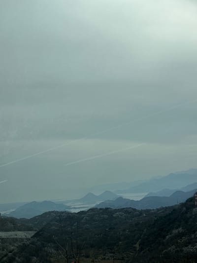 This image presents an expansive landscape vista observed from an elevated position in the area of Gornji Ulići, Montenegro. The scene is dominated by a largely overcast, light grey sky that occupies the upper two-thirds of the frame. Two distinct, parallel white contrails are visible within the sky, traversing diagonally from the upper left quadrant towards the lower right. Below the sky, multiple layers of mountains and hills recede into the distant haze, displaying a gradual blue tonal shift indicative of atmospheric perspective. A large body of water, consistent with a lake or wetland system, is visible horizontally across the midground, situated between the distant mountain ranges. The immediate foreground features dark, densely vegetated hillsides composed primarily of bare or sparsely leafed trees, suggesting a dormant or early growth season. A small, reddish-brown structure is partially visible on a distant hillside at the far right of the image. No individuals, specific actions, or direct interactions are present.