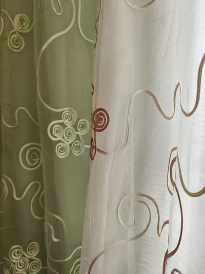 The image shows a close-up view of two sheer curtains. The left curtain is a muted green with embroidered light green swirling and spiral designs. The right curtain is a lighter, off-white color and features embroidered designs in shades of light brown and gold, also in swirling and spiral patterns. The curtains appear to be hanging indoors, possibly in a home, with light filtering through them, creating a soft, textured effect. There are no people visible in the image. The specific location in Rybnitsa, Moldova, cannot be determined from the visual information provided.