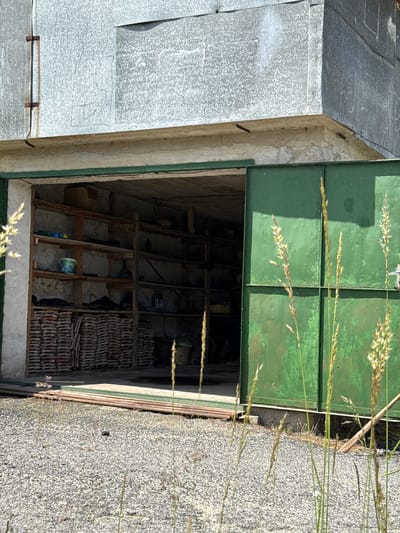 The image depicts a storage area or garage in Ogradna, Bulgaria. The open green door reveals a cluttered interior with wooden framework, stacks of roof tiles, and various containers. To the left of the door is a grey metal structure, likely part of the building's exterior. The surrounding area includes a concrete surface and tall grass. There are no people visible.