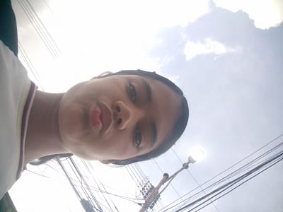 A low-angle, close-up, selfie-style photograph shows the face of a young person looking directly at the camera with pursed lips. The individual is wearing a white t-shirt with a striped collar. The background is a bright, overcast sky with some wispy clouds and numerous overhead electrical wires. A street lamp with its light on is visible in the upper right quadrant of the image, with a pole and tangled wires in the lower right. The overall impression is that the photo was taken outdoors, possibly in a residential area of El Maco, Venezuela, due to the prevalence of overhead electrical wiring, a common sight in many Venezuelan cities. The lighting is diffuse due to the cloud cover.