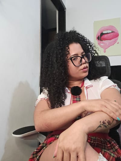 A woman with dark curly hair, wearing glasses, a white top with red trim, and a red plaid skirt is sitting in a black office chair. Her arms are crossed in front of her. She has a floral tattoo on her left arm. Behind her, a painting of lips with a tongue sticking out hangs on the wall. The scene is indoors, in a room with white walls. There is a black object next to the white wall. It is not possible to determine the location in Perímetro Urbano Cúcuta, Colombia, based on the image.
