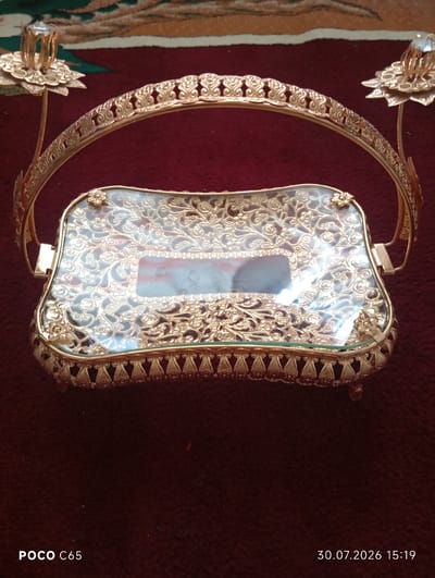 This is a high-angle, medium shot of an ornate golden serving tray with a glass insert, placed on a textured, burgundy rug. The tray features intricate filigree designs, floral motifs, and decorative handles on either side. The glass insert reflects light and shows a blurred image within it. Two crystal-like decorative elements are affixed to the handles. The background is a dark, richly textured rug with a pattern visible in the upper portion of the image. The timestamp "30.07.2026 15:19" and "POCO C65" are visible in the lower portion of the image. There are no people or animals present. The location is identified as Beruniy, Uzbekistan.