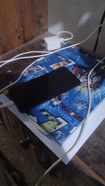 A dark-screened cell phone is plugged into a white charger laying on top of a book. The book has a blue cover featuring Marvel's Avengers characters. The phone and book are sitting on a wooden surface. Location in the city of Putucual, Venezuela cannot be confirmed based on the provided image.