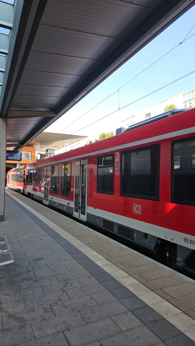 A red and white train is stopped at a station platform in Neu-Ulm, Germany. The train has multiple windows, some of which show passengers inside. The platform is paved with grey stones and has a tactile warning strip along the edge. A modern, covered walkway extends over the platform, with a blue screen displaying train information visible in the background. The sky is clear and blue, and the sun is shining, casting shadows on the platform. The train's exterior displays the "DB" logo and a number "2" on its side. The overall scene depicts a typical urban train station environment.