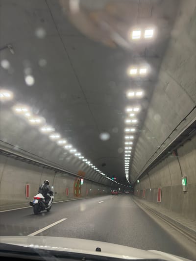 The image depicts the interior of a modern tunnel. The tunnel's ceiling is lined with rows of bright, rectangular lights, illuminating the grey, arched walls. On the left side of the road, a motorcyclist wearing a black leather suit and helmet is riding a white motorcycle with a license plate that reads "P 3392". Further ahead in the tunnel, several cars are visible in the distance, their brake lights faintly glowing. The road surface is asphalt, marked with white lines. Red rectangular panels are mounted on the tunnel walls at intervals. The overall environment is indicative of a well-maintained road infrastructure, likely within a city or major transit route. This scene is specifically located in the tunnel on the A2 highway near the city of Pravets, Bulgaria.