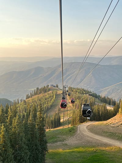 This is an elevated view of the Aspen Mountain Gondola system (Silver Queen Gondola) in Aspen, Colorado, United States. The image is captured from within or near a gondola cabin, looking down the mountainside. Prominent in the foreground is a thick steel cable on the left, with multiple thinner support cables extending diagonally across the upper frame. Several gondola cabins are visible along the cables, including at least two red ones and one black cabin in the immediate foreground, each bearing a white mountain peak logo and the word "ASPEN." A metal lattice gondola tower stands in the mid-ground. The mountain landscape features dense evergreen forests, particularly on the left, and a wide, curving dirt path or ski run descending through areas of green grass on the right. In the distance, layers of mountains recede into a hazy horizon under a clear sky with faint clouds. The town of Aspen is visible nestled at the base of the mountains in the mid-ground. No individuals are discernible.