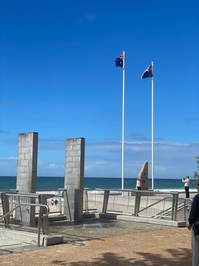 The image captures a section of the Gold Coast, Australia's coastline, near a memorial. Two large, grey, rectangular stone pillars stand prominently in the foreground, with metal railings and a paved pathway leading towards the beach. Beyond these structures, the ocean stretches out under a clear blue sky, with gentle waves breaking on the sandy shore. Two Australian flags fly from tall white poles, one on the left with its flag partially obscured by wind, and another on the right. In the middle ground, a rough, angular stone monument inscribed with "ANZAC" and "LEST WE FORGET" is visible between the flagpoles. A few people are present on the beach and near the memorial area. One individual is walking on the beach with another person, while a man in the mid-right of the frame is seen taking a photograph with a phone, and another person stands near the monument. The overall scene depicts a bright, sunny day at a coastal memorial site.