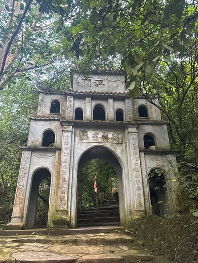 The image shows a weathered, ornate gateway structure in a lush, green forest setting. The gateway, made of gray stone, has multiple arched openings and decorative carvings. It appears to be part of an ancient temple or monument. The structure is adorned with Chinese characters, and there is a flag visible through the central archway. Stone steps lead up to the gateway and continue beyond it into the forest. The overall scene suggests a historical and spiritual site located in a natural environment. The location is in Tây Hoa Lư Ward, Vietnam.