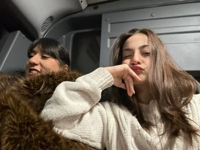 Two young women are in a vehicle, possibly a bus or van. The woman on the left wears a furry brown coat and a white sweater, smiling slightly. The woman on the right, wearing a white sweater, poses with a hand under her chin and a pout. The interior of the vehicle has gray panels and what appears to be a ventilation grill.