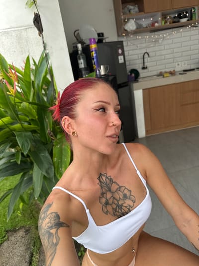 A female individual is depicted from the mid-chest up, positioned in a semi-outdoor living space in Kerobokan, Indonesia. She has bright red hair pulled back, fair skin, and prominent black winged eyeliner. Her left ear displays multiple gold piercings, including helix studs, a hoop, and a large lobe tunnel. Her nose features a double gold hoop piercing on the right nostril, and a small stud is visible below her lower lip. A silver navel piercing is also visible at the bottom of the frame. She wears a white spaghetti-strap crop top. Tattoos include a central chest piece depicting lilies within a geometric pattern, and a large bird tattoo on her left upper arm. Her gaze is directed towards the upper right of the frame, with a relaxed facial expression and slightly parted lips.
To her left and slightly behind, dense tropical foliage with reddish-pink and yellow-tipped blossoms is visible, indicative of an outdoor garden area. The background to her right reveals a modern kitchen setup. This kitchen features light-colored wooden upper and lower cabinets, a light grey countertop, and a white subway tile backsplash. A dark gooseneck faucet is installed at the sink. A dark grey refrigerator stands to the left of the kitchen counter. A string of warm white fairy lights is draped above the backsplash. The floor throughout the visible area consists of large, light grey square tiles.