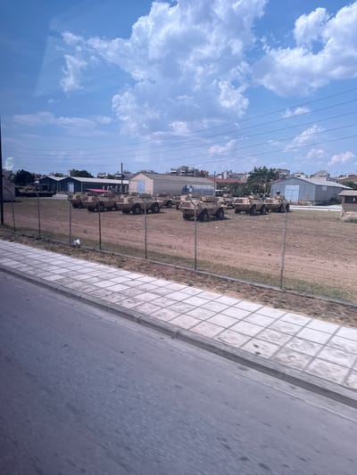 A low-angle, eye-level shot captures a sunny outdoor scene with a clear blue sky dotted with fluffy white clouds. In the foreground, a grey asphalt road runs parallel to a sidewalk paved with light-colored square tiles. Beyond the sidewalk, a chain-link fence encloses a dusty, brown field. Arranged in rows within this field are approximately 15 light-brown armored vehicles, appearing to be military personnel carriers. Behind the field, a row of low-rise buildings, some with white walls and others with grey roofs, forms the urban backdrop. Power lines crisscross the sky above the buildings. The image's perspective suggests it was taken from a moving vehicle, likely a bus or car, looking out through a window, as evidenced by slight reflections on the glass. This scene is located in Stavroupoli, Greece.
