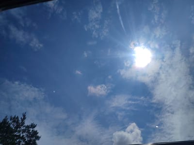 The image shows a bright sunny sky with scattered clouds. The sun is visible in the upper right portion of the frame, with rays of light emanating from it. There is a hint of a dark structure along the top edge, suggesting the photo may have been taken from inside or under a shelter. In the lower left corner, the dark silhouette of a tree with feathery foliage is visible. The overall impression is of a clear, pleasant day. There are no people or specific landmarks visible that would indicate a location in Khorol, Russia, or any other city.