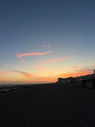The image displays a coastal scene at dusk in Worthing, United Kingdom, with the perspective facing west towards the setting sun. The sky transitions from dark blue at the zenith to pale blue, yellow, orange, and pink hues at the horizon. Several elongated, wispy clouds, predominantly pink, are present in the mid-sky.

The foreground comprises a dark shingle or pebble beach, filling the lower third of the frame. To the left, the dark sea meets an exposed intertidal zone of rocks or sand, consistent with low tide. A single human figure is silhouetted, standing on this exposed intertidal area towards the left-center. Further indistinct human silhouettes are faintly visible along the distant beach.

Along the right side, a row of multi-story buildings, characteristic of a seafront promenade, extends into the distance. Lights from these buildings and streetlights are illuminated, demarcating the urban edge. A structure resembling a beach hut or shelter, featuring an orange-toned interior, is partially visible at the far right edge of the frame. The scene is illuminated by the ambient light of twilight.
