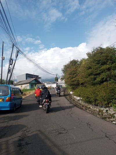 The image depicts a street scene in a mountainous or hilly area. The road is asphalt, cracked in places, and curves slightly to the right. A blue van is on the left side of the road, followed by a yellow vehicle. Several motorcycles are traveling in the same direction, with riders wearing helmets and jackets. Utility poles with multiple wires run along the left side of the road, obscuring a house. A dense row of bushes and a low stone wall line the right side of the road. The sky is blue with scattered clouds. The specific location in Mya Sit Kaing Ywar Thit, Myanmar, cannot be confirmed from the visual details alone.