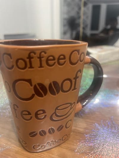 A brown ceramic coffee mug with embossed text and designs sits on a wooden table. The mug is decorated with repeating phrases such as "Coffee Co" and coffee bean motifs. The background is blurred, showing hints of indoor furnishings and lighting. The table surface is partially covered with glittery, festive decorations. The image does not provide any information that would indicate its location in Rosenovo, Bulgaria, or any specific city.