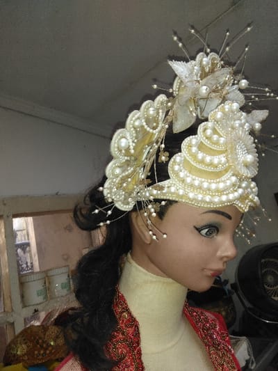 This image displays the head and upper torso of a female mannequin in profile, positioned in an indoor setting. The mannequin has a light brown skin tone, dark hair styled into a long, wavy ponytail, blue-green eyes with dark eyeliner, and light reddish lips. It wears an elaborate, multi-tiered headpiece primarily composed of white and cream faux pearls, gold-toned elements, and translucent white decorative features resembling leaves or butterfly wings, with numerous thin wires tipped with small pearls extending upwards and outwards. A light cream-colored high-neck garment covers the mannequin's neck and upper chest, beneath a red outer garment with intricate gold and red embroidery visible on the right shoulder.
The background on the left shows a wooden-framed opening, possibly a window, with a blurred view of an indistinct light-colored interior or exterior beyond it. Below this, two white plastic containers with green labels are visible on a surface, alongside a textured gold fabric item. To the right, a dark, circular object, possibly a fan, is partially visible. The walls appear light grey or off-white, showing some texture, and the ceiling is a uniform light grey. The specific location within Laguna, Costa Rica cannot be determined from the visual information present in the image.