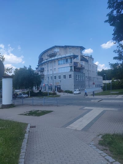 A wide shot captures a modern, curved apartment building with multiple balconies and a sleek, dark roof against a bright blue sky with scattered white clouds. The building has a beige facade and many windows, some with closed white blinds. In front of the building, there is a street with a pedestrian crossing.
On the sidewalk in front of the building, there are a few shops visible, including one with a prominent red sign that appears to be a pharmacy. Text on the building reads "CRILLON" and "FÜR DAS LEBEN". Further down the street, a person in a blue shirt is walking with a wheeled suitcase. A white car is parked near the building, and a silver car is parked further down the street.
The scene appears to be set in a residential or mixed-use urban area during the daytime, likely late morning or early afternoon given the bright sunlight. The weather seems pleasant with a clear sky. The foreground shows a paved walkway and a patch of grass, with a metal railing separating the sidewalk from the street. A tall, white cylindrical object, possibly a lamp post or informational pillar, stands on the left side of the frame. Trees line the background, suggesting a somewhat green environment within the urban setting. The overall impression is one of everyday urban life on a sunny day.