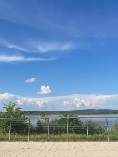 The image depicts a view from a paved area in Shiryaevo, Russia. In the foreground, a paved surface with rectangular tiles is visible. A metallic railing with horizontal bars and vertical posts separates the paved area from the natural environment. Behind the railing, lush green grass and various trees and bushes are present, forming a dense foliage. Beyond the vegetation, a body of water, likely a river, stretches across the midground, with a distant, tree-covered hillside forming the horizon. The sky above is a vibrant blue, adorned with scattered white cumulus clouds. The scene appears to be a serene outdoor location, possibly a park or a viewpoint, offering a picturesque landscape.