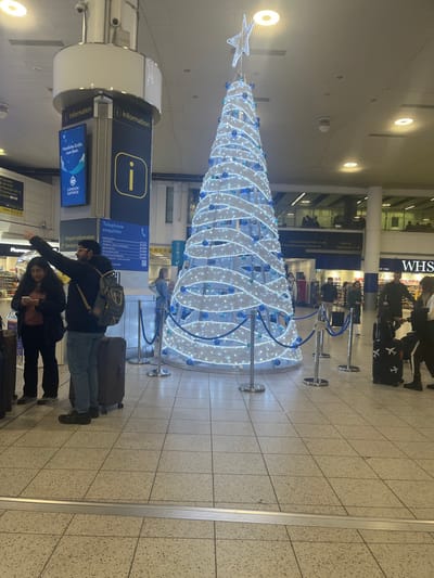 The image shows an indoor public space, likely an airport terminal or large train station, decorated for the Christmas season. The main subject is a large, tall, conical Christmas tree illuminated with white lights and adorned with blue ornaments or ribbons, topped with a white star. The tree is surrounded by silver queue barriers.
In the foreground, two individuals are visible. A man, wearing a backpack and holding a suitcase, is pointing towards something out of frame to the left. Beside him, a woman with long dark hair is holding a mobile phone and also appears to have luggage. Both are standing near a circular information kiosk.
The information kiosk features a prominent "i" symbol and text that reads "Information," "Telephone enquiries," and "Airport Information Service." One of the screens on the kiosk partially displays text including "London Gatwick," indicating the location is likely London Gatwick Airport.
In the background, more queue barriers are visible, leading towards what appears to be a shopfront with the letters "WHS" (likely WHSmith, a common retailer in UK transport hubs). Other blurry figures of people can be seen in the distance near the shop. The floor is tiled, and the ceiling features recessed lighting. The overall scene suggests a busy travel environment during the holiday period.