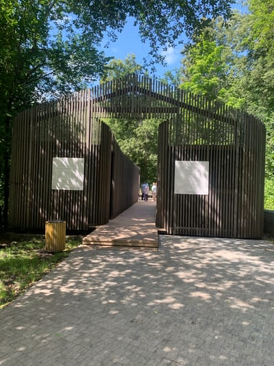 The media file captures a view of a wooden structure with horizontal slats, likely a part of a walkway or an architectural installation, in a natural setting. Tall green trees with lush foliage surround the structure, and the sky above is a clear blue with a few wispy clouds. Sunlight filters through the leaves, casting dappled shadows on the textured, light-colored paving stones that form a path alongside the wooden structure.

The scene appears to be outdoors, possibly in a park or a wooded area. The time of day seems to be daytime, with bright sunlight indicating it is not early morning or late evening. The weather appears to be fair and sunny.

In the distance, within the walkway, several people are visible, appearing to be walking or conversing. They are dressed casually, suggesting a relaxed atmosphere. One of the notable details is a white plaque mounted on the wooden structure, bearing text that is likely informative about the location or the structure itself. There is also a cylindrical wooden bin next to the structure.

The overall impression is one of a tranquil outdoor space designed for visitors to explore and appreciate nature, possibly with an artistic or historical element. The contrast between the dark wood and the bright paving stones, along with the interplay of light and shadow, adds visual interest to the scene. The orientation of the image is vertical.