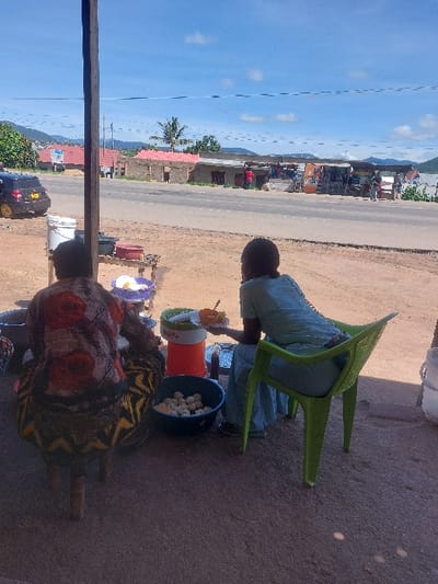 The scene depicts an outdoor roadside area in Kimamba, Tanzania, under a bright blue sky with scattered white clouds. In the immediate foreground, two individuals are visible from the back. On the left, a person wearing a multi-colored floral-patterned shirt and a patterned skirt is seated on a low, three-legged wooden stool. Adjacent to this person is a dark blue basin containing numerous white, spherical food items. A small wooden table holds a white bucket, a dark rectangular tray, a small bowl with white food, an orange and white insulated beverage dispenser, and a small dark bottle. On the right, a second person, dressed in light blue attire, is seated on a lime-green plastic chair, holding an orange bowl with food and a spoon, and a small white plate positioned over a metal tray. A thick wooden pole stands vertically in the left foreground, partially obscuring the view.
A paved road spans the midground. Across the road, a series of single-story structures includes an unfinished brick building with a red roof, alongside several small shops displaying various goods. Multiple individuals are visible near these shops. A black vehicle with yellow accents on its wheels is parked on the left side of the road. Overhead utility lines traverse the midground. In the distant background, green hills are visible against the horizon. The area is illuminated by bright daylight, casting visible shadows.