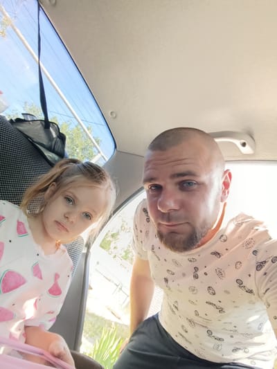 This is a selfie taken inside a vehicle, likely a car, from a low angle looking up. The primary subjects are a man and a young girl. The man, in the foreground and to the right, is bald with blue eyes and a beard. He is wearing a white t-shirt with a repeating pattern of black motorcycles and gears. The girl is to the left and slightly behind the man. She has blonde hair tied in two ponytails and striking blue eyes. She is wearing a light-colored t-shirt with a pattern of pink watermelons. The background shows the interior of the car, including the back seats, a car handle, and the rear window. Through the window, trees and a blue sky with some sunlight are visible, suggesting it is daytime and likely sunny weather. The overall mood appears casual and perhaps a bit candid. The scene is set within a vehicle, indicating travel or an outing. The location context provided is Pishchane, Ukraine. There is no visible text in the image.